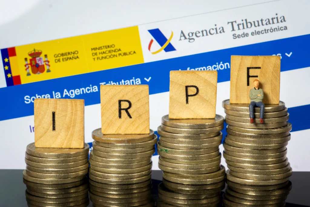Bloques con letras IRPF sobre pilas de monedas junto al logo de la Agencia Tributaria