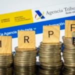 Bloques con letras IRPF sobre pilas de monedas junto al logo de la Agencia Tributaria
